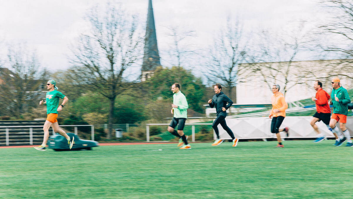 Ein Lauftrainer läuft voran, eine Gruppe aus Läuferinnen und Läufern joggt hinterher. Offensichtlich ein Firmenlaufkurs. Geführte Laufkurse machen aus einem Firmenlauf eine betriebliche Gesundheitsmaßnahme.